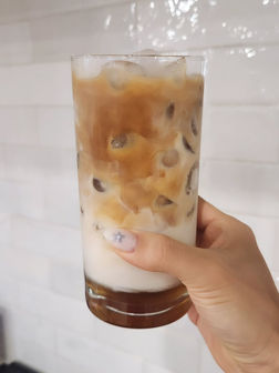 Hand holding iced coffee in a tall glass, white tile background.